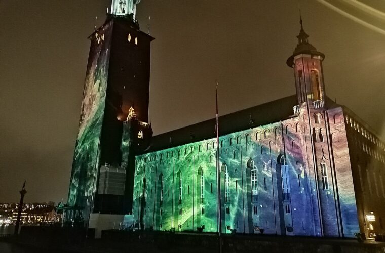 Stockholm City Hall at night, its brick tower and long facade washed in green, blue and violet projected light during Nobel Week Lights