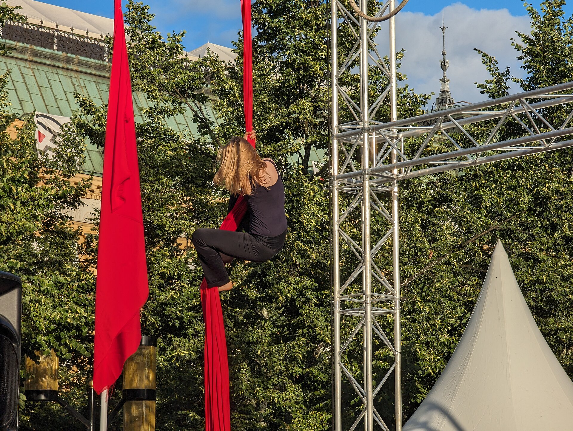 An aerial performer climbs a long red silk hanging from a metal rig in front of summer trees and a copper roof at the Stockholm Culture Festival