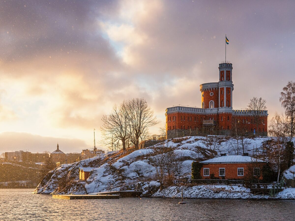 The red-brick Kastellet citadel with its flag tower on snow-covered rocks of Kastellholmen, lit gold by a low winter sun as snowflakes fall over the water