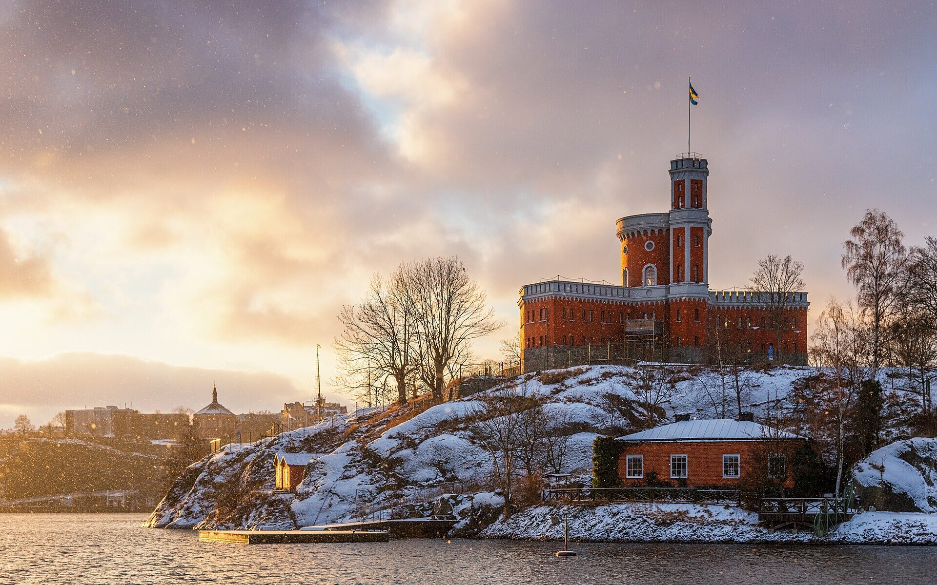 The red-brick Kastellet citadel with its flag tower on snow-covered rocks of Kastellholmen, lit gold by a low winter sun as snowflakes fall over the water