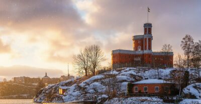 The red-brick Kastellet citadel with its flag tower on snow-covered rocks of Kastellholmen, lit gold by a low winter sun as snowflakes fall over the water