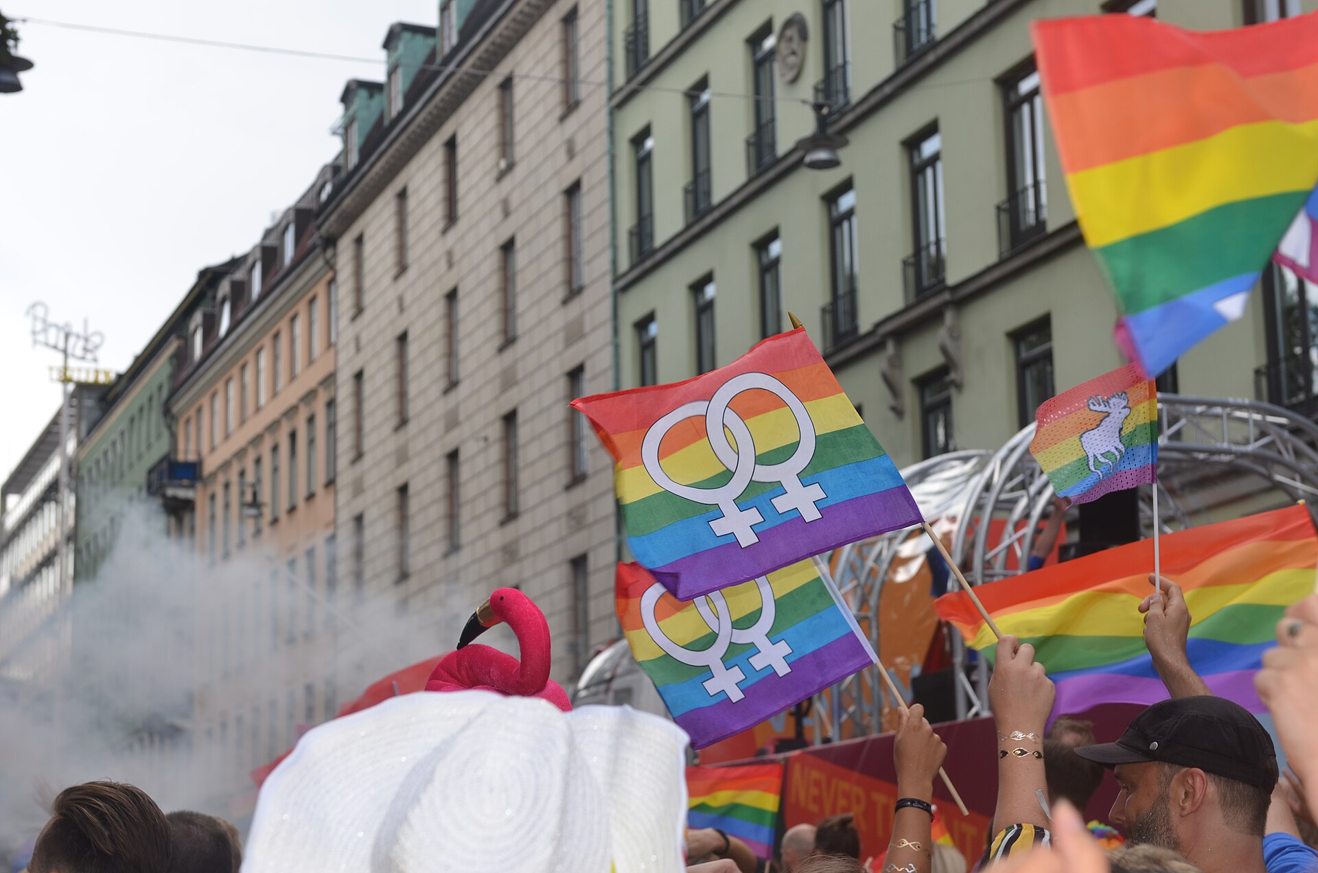 Rainbow flags and flags with linked Venus symbols held high above a Pride parade crowd between tall Stockholm buildings