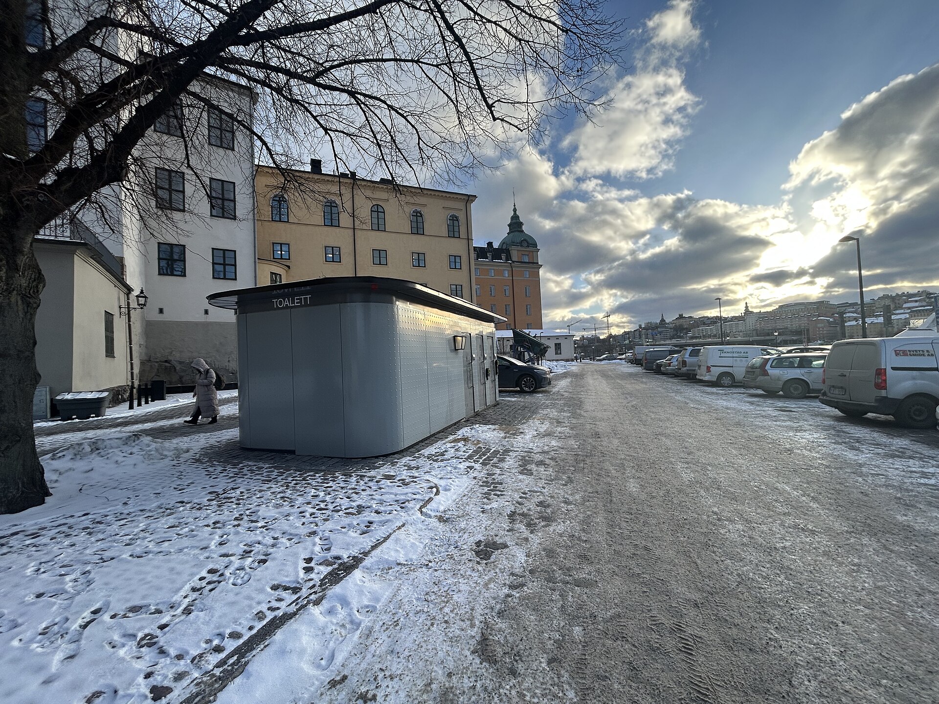 A grey modular building marked TOALETT on the quayside at Södra Riddarholmshamnen, with snow on the ground and parked cars along the water