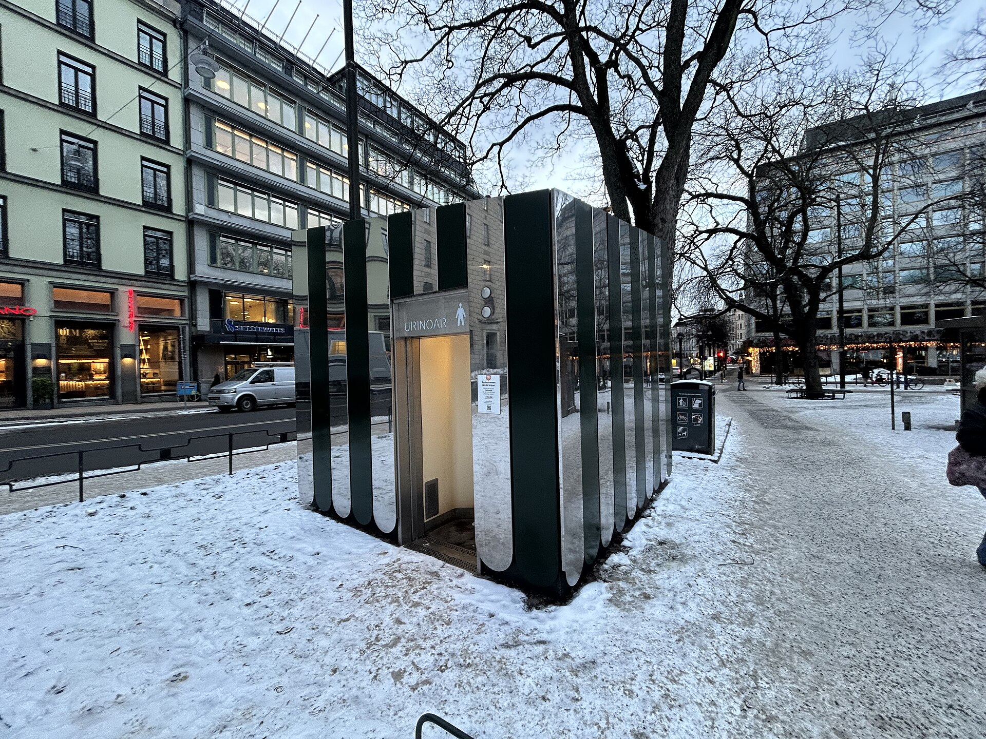 A mirrored stainless-steel self-cleaning public toilet cabin on a snowy pavement beside Humlegården, with city buildings behind