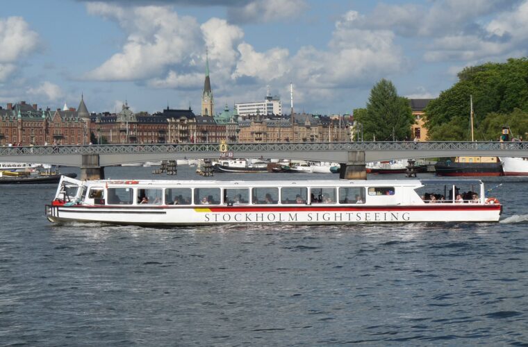 A long white Stockholm Sightseeing boat full of passengers cruising past Djurgårdsbron and the grand facades of Strandvägen under a cloudy summer sky
