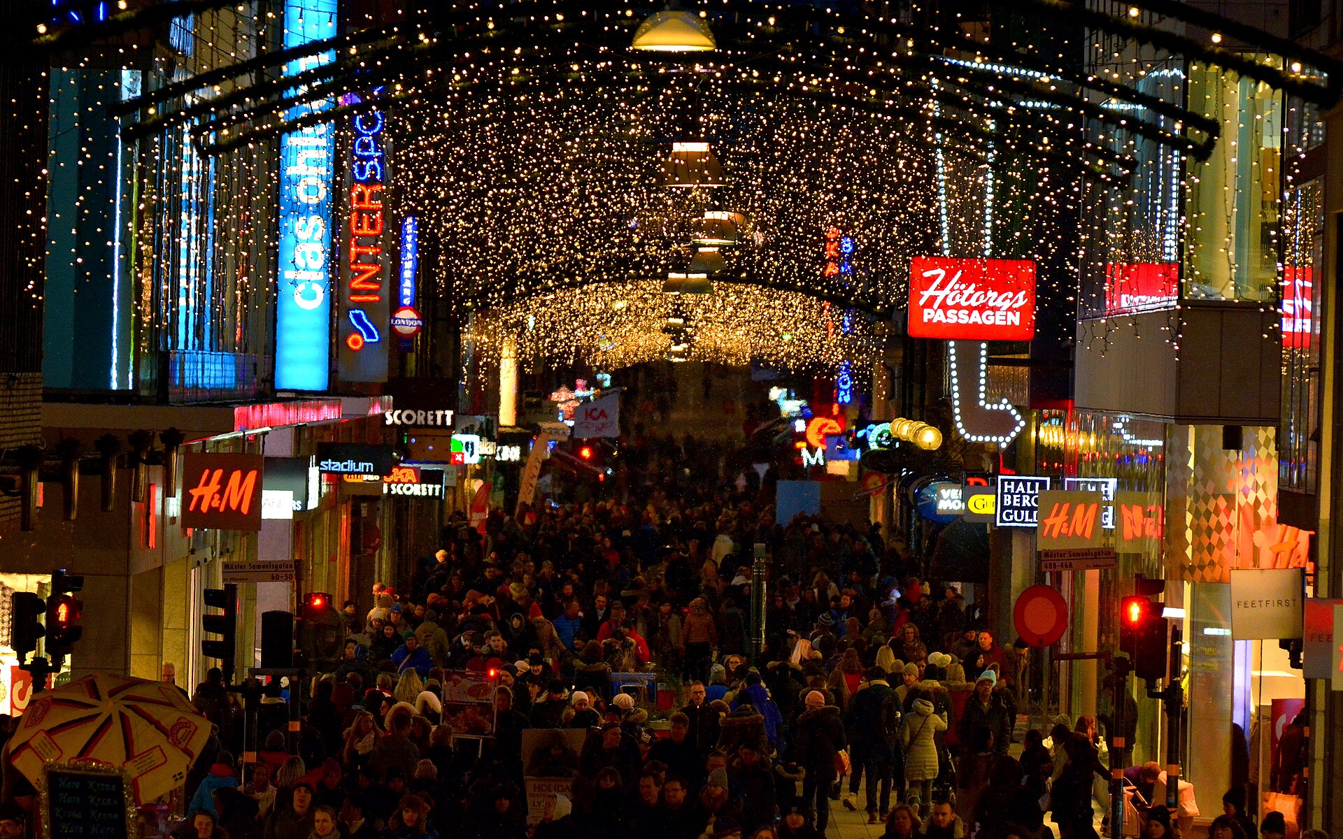 Dense evening crowds under Christmas lights on Drottninggatan in Stockholm, with illuminated shop signs
