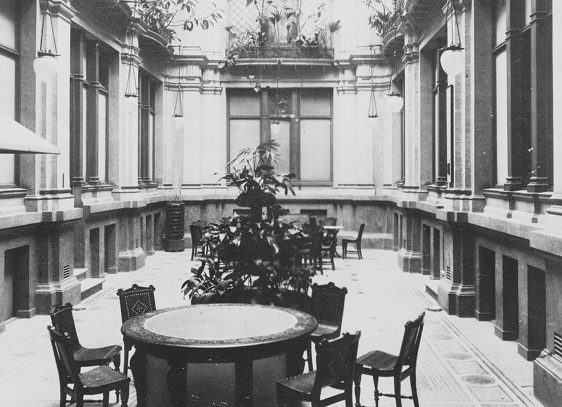 Black-and-white photograph of Sturebadet's atrium in 1896, a tall marble hall with hanging globe lamps, potted palms and café tables and chairs