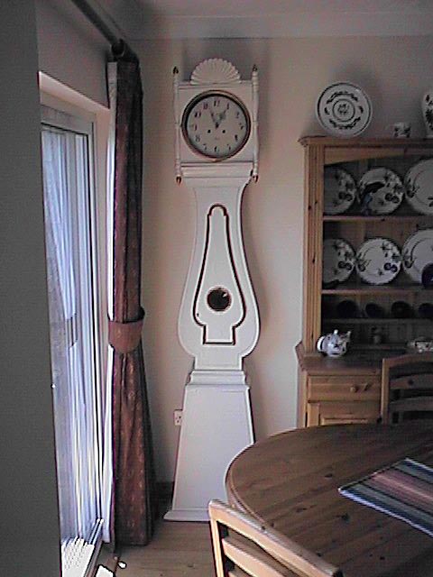 A tall white-painted Mora longcase clock with a curved waist standing in a Swedish dining room beside a dresser of blue-and-white plates