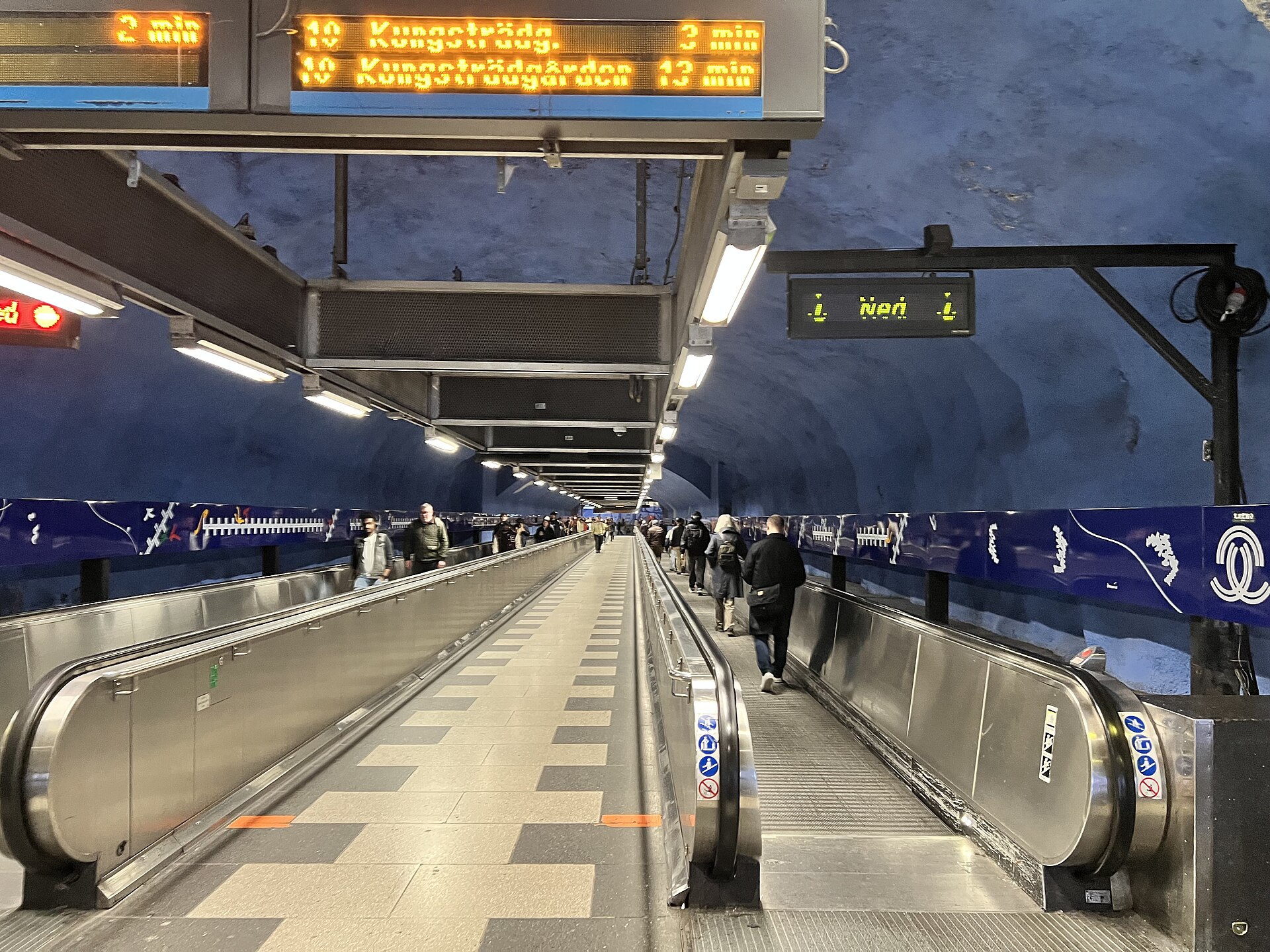 A long moving walkway and escalators under the blue painted ceiling of the passage between platforms at T-Centralen