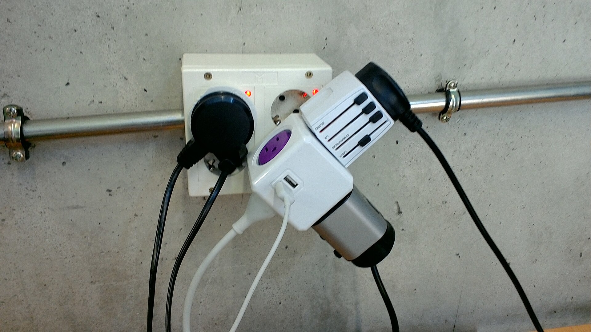 A universal travel adapter with USB ports plugged into a wall-mounted Schuko socket alongside another plug, with several cables attached